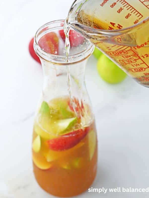 Apple cider punch being poured from a glass measuring cup into a carafe with apple slices and ice. 