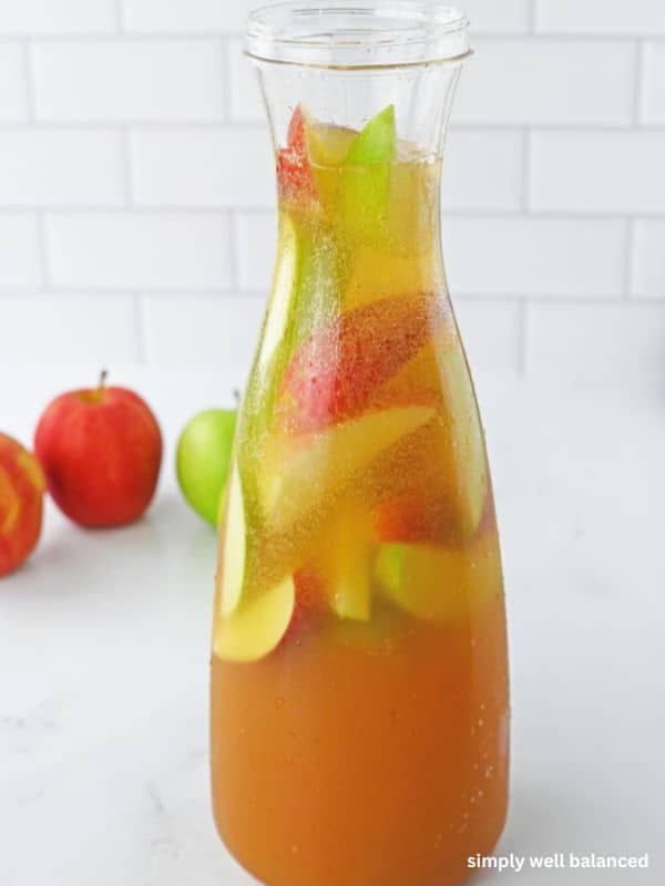 A tall glass carafe filled with fall Apple Pie Punch and apple slices. 