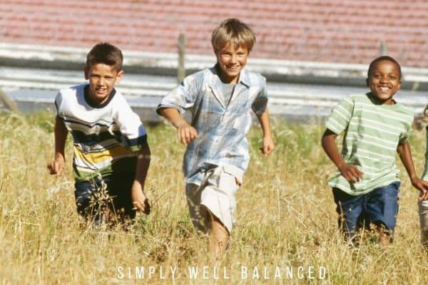 Three young, smiling boys run around in a field of overgrown grass.