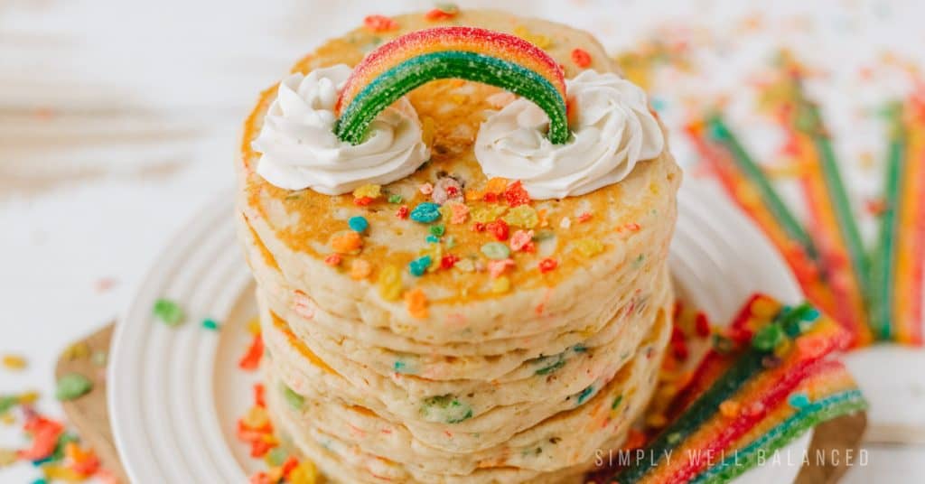 A stack of colorful pancakes made with Fruity Pebble Cereal and topped with a candy belt rainbow and whipped cream clouds. 