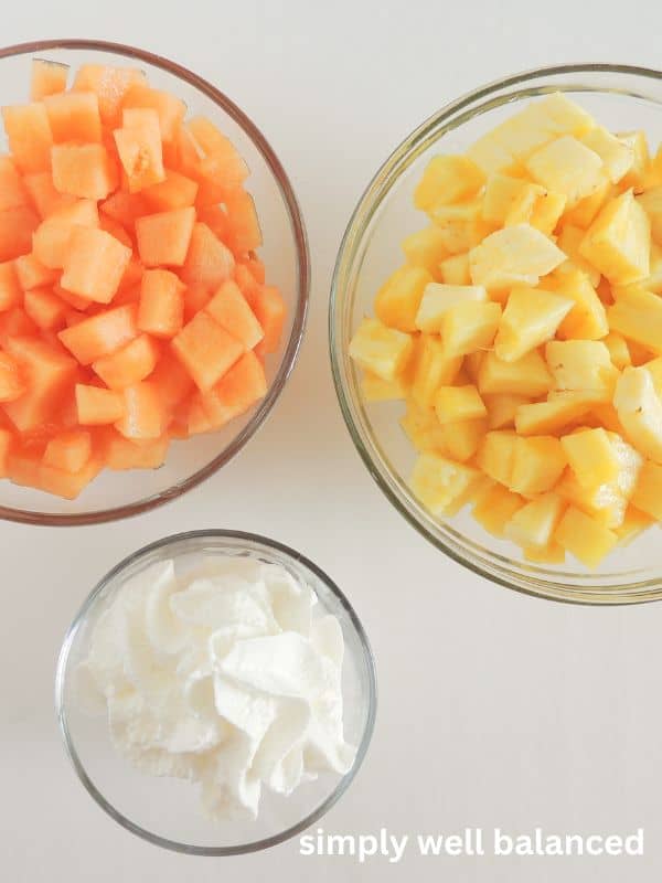 Ingredients for candy corn fruit parfaits: diced pineapple in a glass bowl, diced cantaloupe in a glass bowl, and whipped cream.