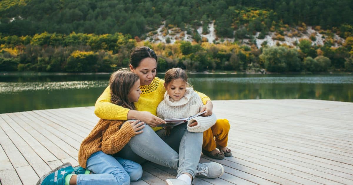 Mom spending time with two daughters reading a book on a dock overlooking a lake.